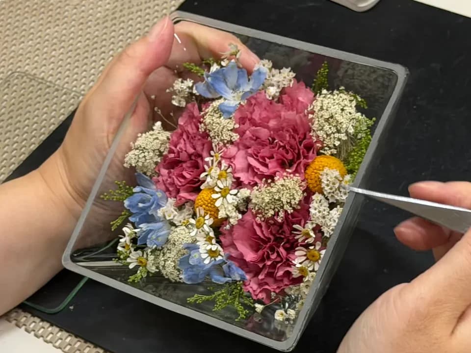 ガラスケースに収めたピンクのカーネーションや青い花をピンセットで整える手元のアップ